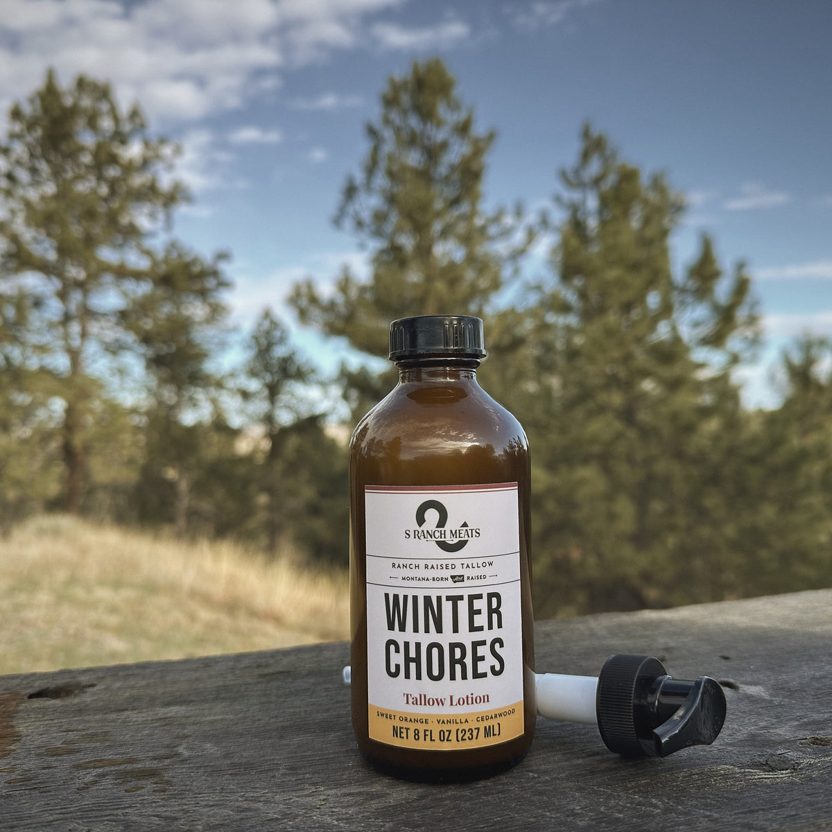 Winter Chores tallow lotion, 8 oz amber bottle with pump, on a ranch table under Montana pines