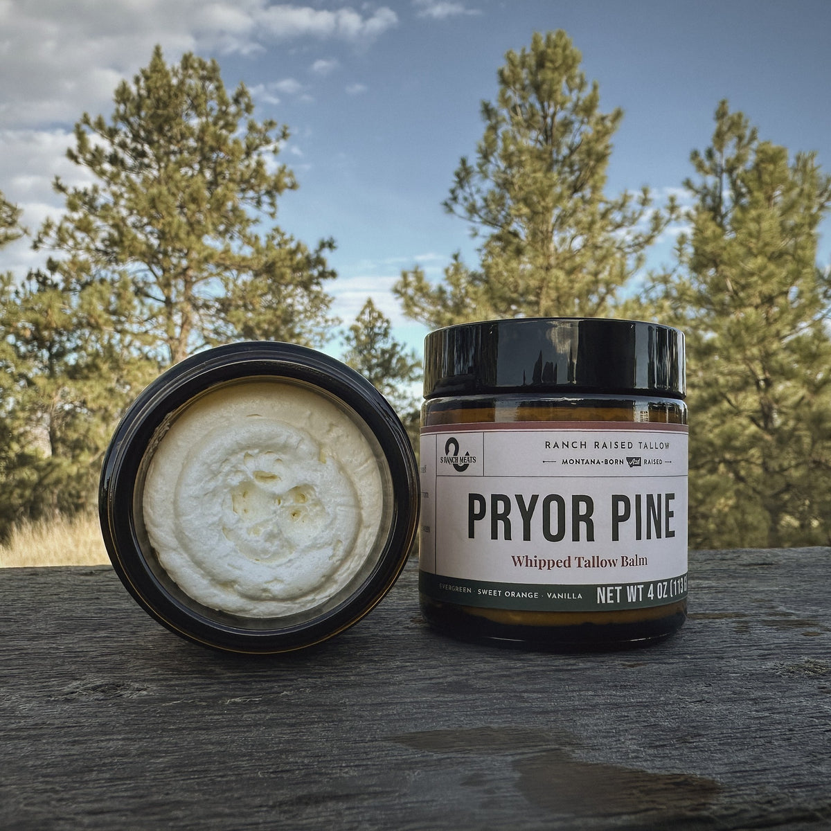 Pryor Pine whipped tallow balm, 4 oz amber jar with lid off, on a weathered ranch table under Montana pines